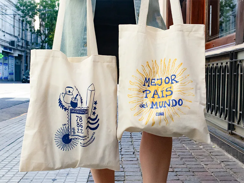 Dos tote bags de lienzo crudo estampadas en azul y amarillo, en una vereda de Buenos Aires